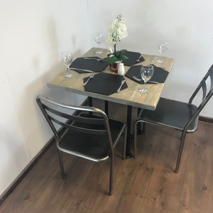 May include: A square wooden table with a distressed finish, set for four with black placemats, cutlery, and glasses. The table is surrounded by four black metal chairs with a simple design.