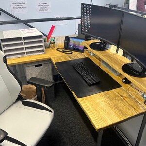 May include: A corner desk with a light wood finish and black metal legs. The desk has a black mouse mat and a keyboard on it. There are two computer monitors on a shelf above the desk. A white office chair is in the foreground.