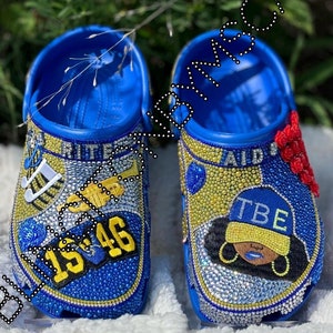 May include: A pair of blue and yellow custom-designed clogs with rhinestones. One clog features a musical note design and the text "1546". The other clog features a black and white portrait of a woman wearing a blue and yellow hat with the text "TBE".