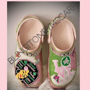 May include: Pink and green embellished clogs with a pink and green 'LOVE' design on one clog and a pink and green 'AKA' design on the other clog. The 'AKA' design includes a black and pink illustration of a woman's face with the text 'Strong Sisterhood, Leader, Educated, Gifted, Brave, Pretty, Blessed, Proud, Loving, Pride'.