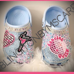 May include: A pair of light blue clogs decorated with rhinestones and colorful patches. One clog features a pink ballerina, a pink heart with a silver glitter design, and a pink and silver disco ball. The other clog features a pink disco ball with the words "dancing queen" in pink.