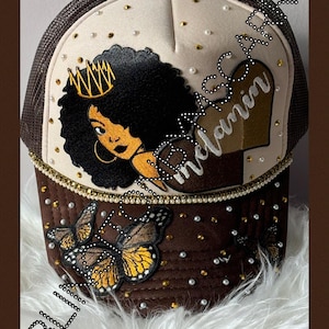 Puede incluir: Gorra de camionero marrón y beige con la parte trasera de malla negra. La parte delantera presenta una ilustración de una mujer negra con una corona dorada, la palabra "melanina" y un diseño de mariposa. Adornada con perlas y tachuelas doradas.