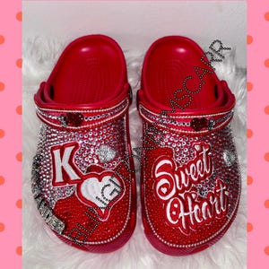 May include: Red clogs with rhinestones and embroidered designs. One clog says "K" with a heart and the other says "Sweet Heart".