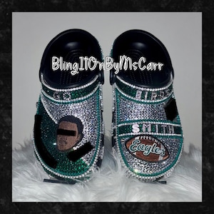 May include: Pair of black Crocs customized with green and silver rhinestones. One shoe features a portrait and the other has a football graphic with the text "Eagles". The text "Smith" is also visible. The text "BlingITOnByMsCarr" is also visible.