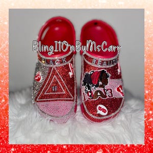 May include: A pair of red Crocs adorned with sparkling rhinestones and decorative embellishments. One shoe features a triangle design, while the other showcases a graphic of a woman. The text "Bling It On By MsCarr" is visible.