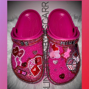 May include: A pair of pink clogs decorated with rhinestones and Valentine's Day themed embellishments. The clogs have the words "Be Mine" on one and "Love you" on the other.
