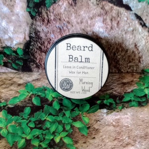 May include: A round tin container with a black lid and a label that reads "Beard Balm Leave in Conditioner Wax for Men Morning Wood Net Wt. 1 oz."