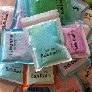 May include: Assortment of colorful bath dust packets in various scents. The packets are sealed in clear plastic with labels that read "Bath Dust" and scent names like "Gummy Bears" and "Lavender & Chamomile". The packets are in shades of blue, green, pink, and purple.