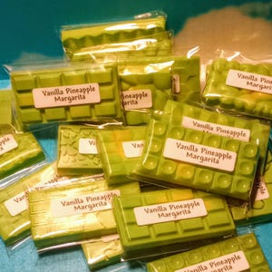 May include: A close-up of multiple green wax melts, individually packaged in clear plastic bags. The wax melts are all scented with "Vanilla Pineapple Margarita".
