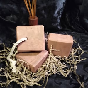 May include: Three square, light brown soaps with a rope detail, arranged on brown paper filler. A small wooden container with several brown sticks is in the background. The background is a dark, patterned fabric.