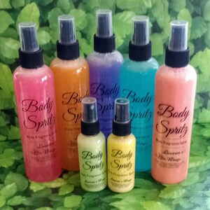 K&ouml;nnte beinhalten: Sieben Glasflaschen mit schwarzen Spr&uuml;hk&ouml;pfen und Etiketten, auf denen "Body Spritz" in einer kursivschriftlichen Schriftart steht. Die Flaschen sind mit verschiedenen farbigen Fl&uuml;ssigkeiten gef&uuml;llt: rosa, orange, lila, blau und pfirsich. Die Etiketten tragen au&szlig;erdem die Aufschrift "Body Fragrance Spray" und "Lauren's Wings".