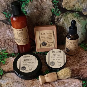 May include: A set of grooming products for men, including beard wash, beard oil, beard balm, shave soap, and man soap. The products are all in brown glass bottles and jars with black labels and the text "Morning Wood" on them. The products are arranged on a brown wooden surface.