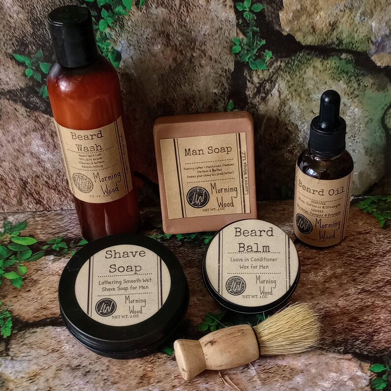 May include: A set of grooming products for men, including beard wash, beard oil, beard balm, shave soap, and man soap. The products are all in brown glass bottles and jars with black labels and the text "Morning Wood" on them. The products are arranged on a brown wooden surface.