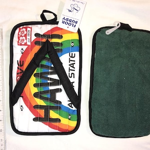 Puede incluir: Dos agarraderas rectangulares. Una presenta un fondo blanco con un diseño de arcoíris y la palabra "HAWAII" en negro. La otra es verde. Ambas tienen ribetes negros y un lazo para colgar. La etiqueta dice "FLOOR BUDDY".