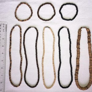 May include: A selection of necklaces and bracelets crafted from natural materials. The necklaces range in colour from light beige to dark brown and black. The bracelets are circular. A ruler is placed next to the necklaces.
