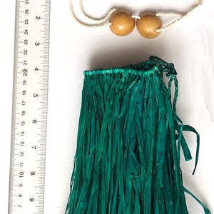 Puede incluir: Una falda de hierba verde y un collar de cuentas de madera. La falda está hecha de tiras largas y finas de material verde. El collar tiene dos cuentas redondas de color marrón claro. La falda mide unos 20 cm.