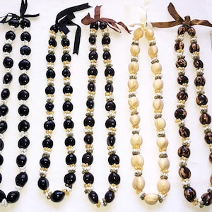 May include: Five strands of necklaces with varying designs. The necklaces feature large, dark beads, light-colored beads, and smaller accent beads. Each necklace is tied with a ribbon at the top. A ruler is placed next to the necklaces.