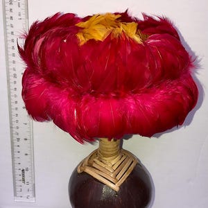 Pode incluir: Objeto decorativo com uma parte superior de penas vermelhas vibrantes e detalhes amarelos. É montado sobre uma base arredondada castanha escura com detalhes de palha tecida. O objeto tem cerca de 25 cm de altura.