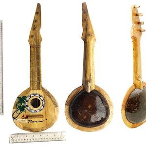 May include: Three wooden ukulele-style instruments. The instruments have a light brown wooden body with a dark brown coconut shell base. One ukulele has a painted scene with palm trees and the word "Hawaii". A ruler is next to the instruments.
