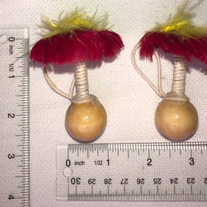 Pode incluir: Dois objetos decorativos em forma de cabaça. Cada um tem uma base esférica marrom clara, um caule enrolado e um topo de penas vermelhas com detalhes amarelos. Uma corda fina e curva se estende do caule à base. Objetos em uma superfície branca.