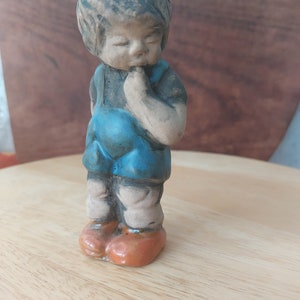 Może przedstawiać: Ceramiczna figurka małego chłopca z brązowymi włosami, ubrana w niebieski kombinezon i pomarańczowe buty. Chłopiec patrzy w dół i ma rękę na brodzie, jakby się zastanawiał.
