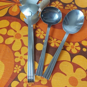 May include: Three silver kitchen utensils on a patterned orange and yellow background. The utensils include a serving spoon, a slotted spoon, and a salad server.