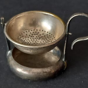 Könnte beinhalten: Ein Teesieb aus silberfarbenem Metall mit einer perforierten Schale, einem geschwungenen Griff und einem runden Standfuß. Das Sieb hat eine polierte Oberfläche mit sichtbaren Gebrauchsspuren und Oxidation und ist zum Abstellen konzipiert.