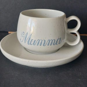 Peut inclure: Un ensemble tasse et soucoupe en céramique blanche mouchetée. La tasse a une forme arrondie avec une anse et le mot "Mumma" en écriture bleue. La soucoupe est ronde et correspond au design de la tasse.