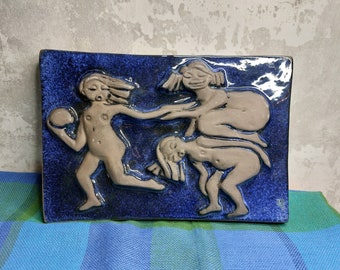 May include: A ceramic wall tile with a blue background and three figures in relief. The figures are nude and appear to be dancing or playing. The tile has a glossy finish.