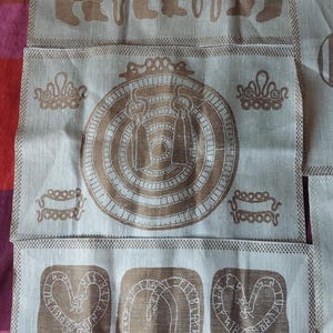 May include: A set of three beige linen tea towels with brown embroidered designs. The first towel features three figures with long staffs, the second towel has a circular design with concentric circles, and the third towel has three stylized figures with runes.