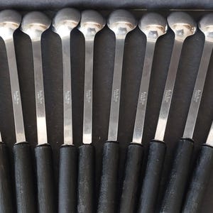 May include: A collection of silver spoons with black handles. The spoons have a rounded bowl and a long, slender handle. The handles are black and appear to be made of a rubberized material. The spoons are arranged in a row on a dark surface.