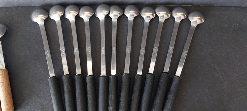May include: A collection of silver spoons with black handles. The spoons have a rounded bowl and a long, slender handle. The handles are black and appear to be made of a rubberized material. The spoons are arranged in a row on a dark surface.