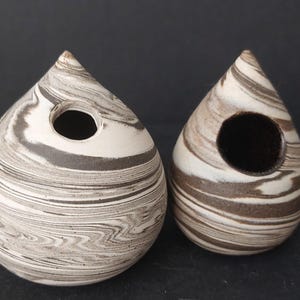 Pode incluir: Duas casas de pássaros de cerâmica em forma de gota com um padrão marmorizado em tons de branco e castanho. Cada casa de pássaros tem uma abertura circular. As casas de pássaros são exibidas em uma superfície escura.