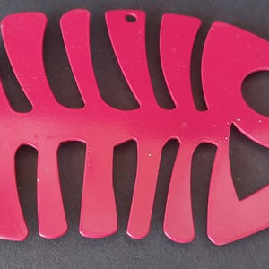 Puede incluir: Un salvamanteles de cocina de metal rojo brillante con forma de esqueleto de pez. El salvamanteles tiene un agujero en la parte superior para colgarlo y está diseñado para proteger las superficies de los utensilios de cocina calientes. El diseño es una representación estilizada de la estructura interna de un pez.