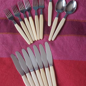 Può includere: Una collezione di posate vintage, tra cui forchette, cucchiai, coltelli e un mestolo, disposte su una tovaglia a righe rosse e viola. Le posate presentano manici color crema e teste in acciaio inossidabile.