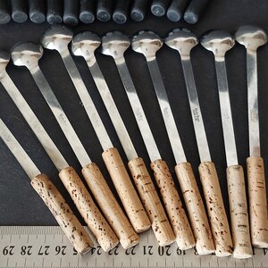 May include: A collection of silver spoons with cork handles. The spoons have a round, flat head and a long, slender stem. The cork handles are cylindrical. The spoons are arranged on a dark surface.