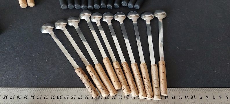 May include: A collection of silver spoons with cork handles. The spoons have a round, flat head and a long, slender stem. The cork handles are cylindrical. The spoons are arranged on a dark surface.