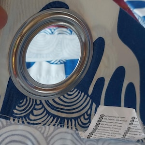 Puede incluir: Primer plano de un panel de cortina con una arandela plateada. La tela presenta un fondo blanco con un patr&oacute;n de olas azules. Una etiqueta con texto es visible en la parte inferior derecha. La cortina est&aacute; hecha de una mezcla de poli&eacute;ster y algod&oacute;n.