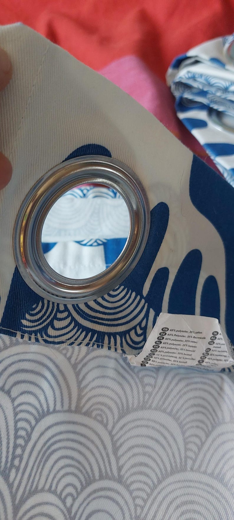 Puede incluir: Primer plano de un panel de cortina con una arandela plateada. La tela presenta un fondo blanco con un patr&oacute;n de olas azules. Una etiqueta con texto es visible en la parte inferior derecha. La cortina est&aacute; hecha de una mezcla de poli&eacute;ster y algod&oacute;n.