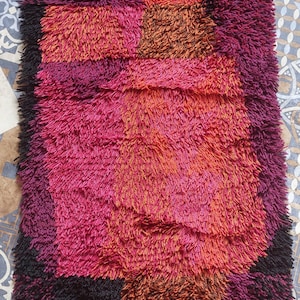 May include: A colorful, abstract rug with a geometric design in shades of pink, orange, and black. The rug has a shaggy texture and is made of wool.