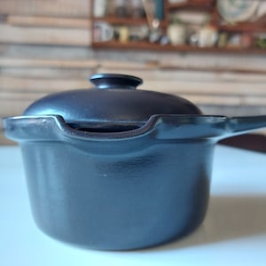 Puede incluir: Una olla de hierro fundido negra con tapa y asa. La olla está sobre una superficie blanca.