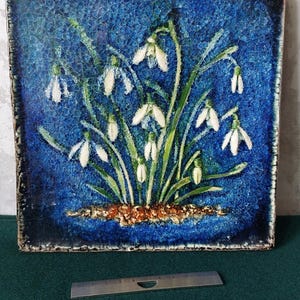 May include: Square ceramic tile with a blue background and a painted image of snowdrop flowers. The flowers are white with green stems and leaves. The tile has a dark border and a string for hanging.