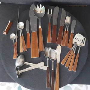 Puede incluir: Una colección de cubiertos de acero inoxidable con mangos de madera, que incluye cuchillos, tenedores, cucharas y utensilios para servir. Los mangos de madera son de color marrón cálido y el metal es brillante. Los cubiertos están dispuestos sobre una superficie gris oscuro.