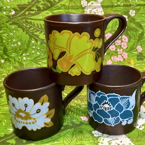 Puede incluir: Tres tazas de cerámica marrón con diseños florales. Las tazas tienen diferentes patrones florales en tonos de amarillo, azul y blanco.