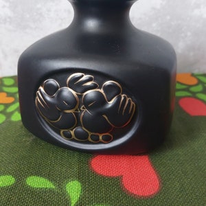 Puede incluir: Jarrón de cerámica negro con un diseño dorado de dos pájaros sosteniendo un corazón. El jarrón tiene una boca ancha y un cuerpo redondeado.