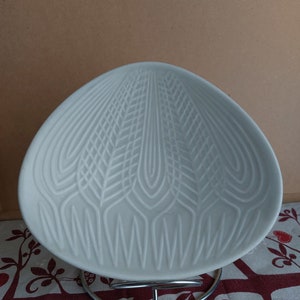 May include: A white, shell-shaped ceramic dish with a detailed, textured pattern. The dish sits on a chrome wire stand. The background is a neutral colour.
