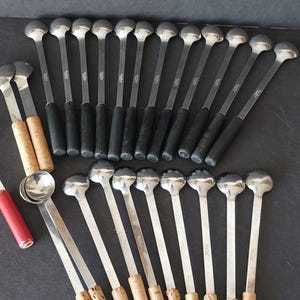 May include: A collection of vintage bar spoons with silver heads and long stems. Some spoons have black handles, while others have cork handles. A red and silver item is also visible.