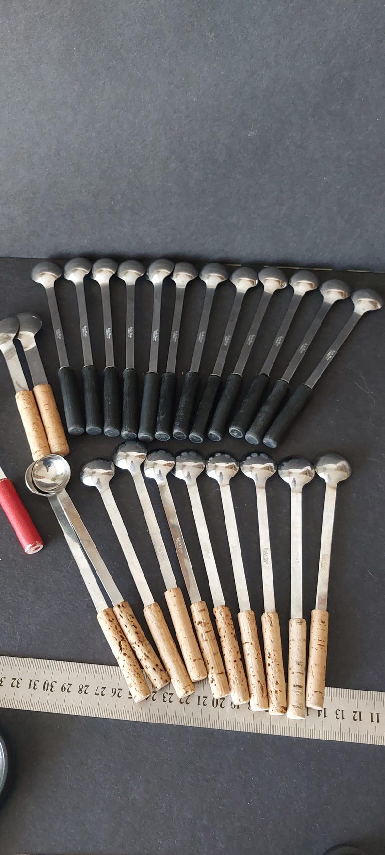 May include: A collection of vintage bar spoons with silver heads and long stems. Some spoons have black handles, while others have cork handles. A red and silver item is also visible.