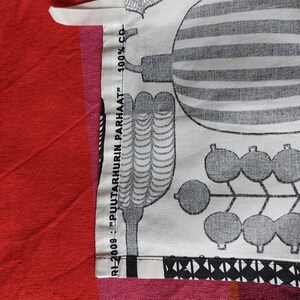 May include: Close-up of a white fabric with a black and white pattern of stylized plants and berries. The fabric has a label that reads "RH-2009... "PULTARHURIN PARHAAT"... 100% CB."