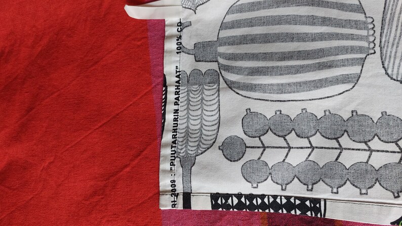 May include: Close-up of a white fabric with a black and white pattern of stylized plants and berries. The fabric has a label that reads "RH-2009... "PULTARHURIN PARHAAT"... 100% CB."